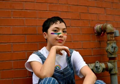 Photo d’un garçon au visage peint aux couleurs de l’arc-en-ciel. Il est assis contre un mur de brique. Illustration du chapitre 1 de l’émission : « Qu’est-ce qu’il se passe dans la tête des homos ? », du podcast Méta de Choc. Ce chapitre s’intitule : Dans la sphère privée.