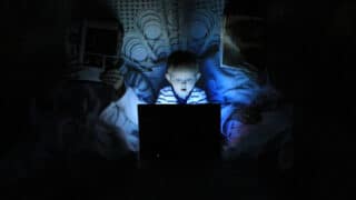 Photo d’un petit garçon jouant dans le noir sur un ordinateur portable dans le lit de ses parents. Illustration du teaser de l’émission : Les algorithmes tueront-ils notre attention ?, du podcast Méta de Choc.