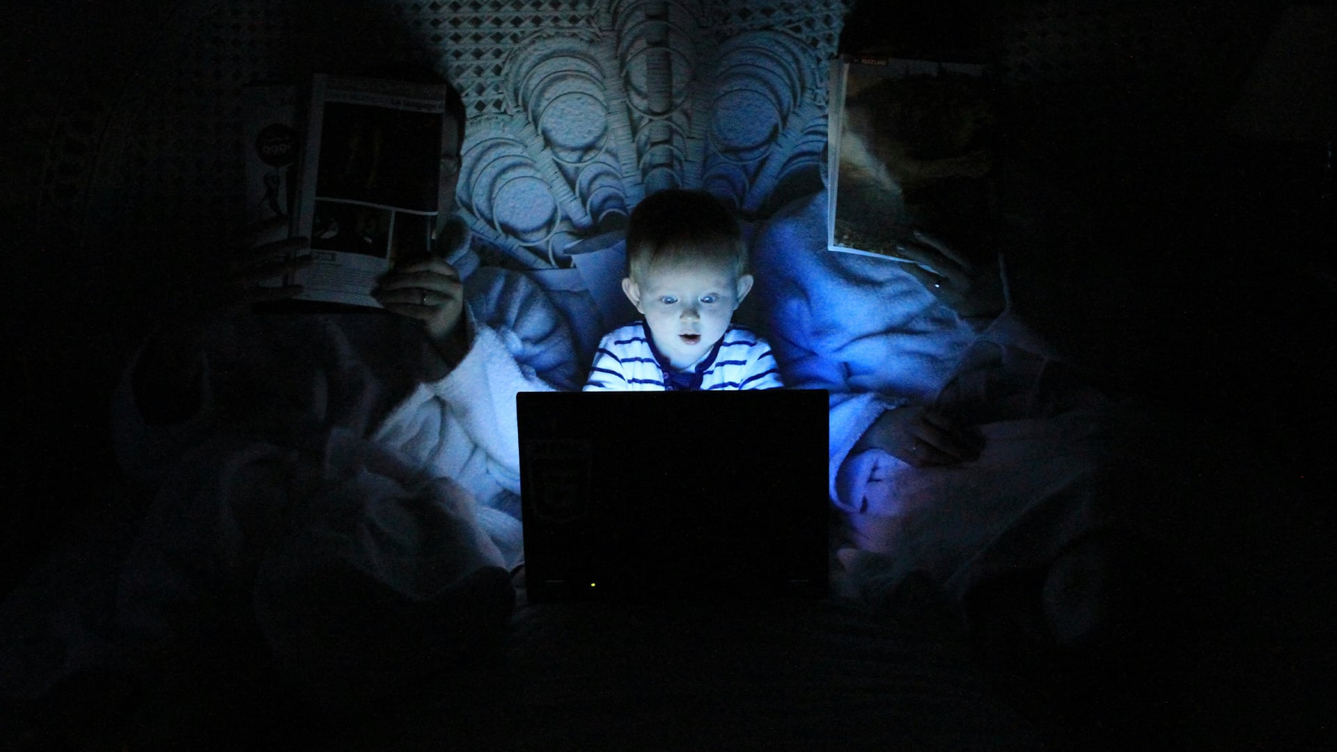 Photo d’un petit garçon jouant dans le noir sur un ordinateur portable dans le lit de ses parents. Illustration du teaser de l’émission : Les algorithmes tueront-ils notre attention ?, du podcast Méta de Choc.