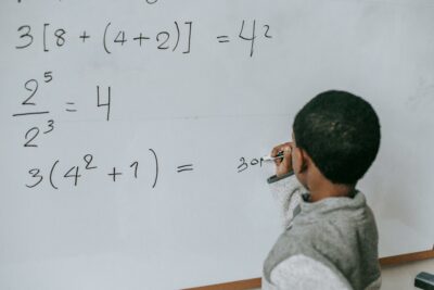 Photo d’un jeune enfant au tableau en train de faire des maths. Illustration du chapitre 1 de l’émission : L’enseignement à la loupe, du podcast Méta de Choc. Ce chapitre s’intitule : La jeune recherche en didactique.