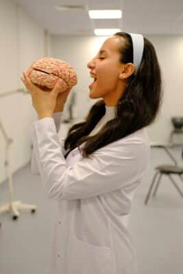 Photo d'une médecin qui tient un faux cerveau dans les mains. Elle fait semblant de le manger. Illustration du chapitre 1 de l’émission : Émotion et rationalité, du podcast Méta de Choc. Ce chapitre s'intitule : La psychologie et les neurosciences sont-elles fiables ?