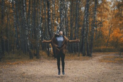 Photo d’une femme au milieu d’une forêt aux couleurs automnales. Elle est en train de sauter, la tête dirigée vers le ciel et les yeux fermés. Illustration de la partie 2 de l’émission : L’écospiritualité, du podcast Méta de Choc. Cette partie 2 s’intitule : Les implications politiques et sociales.