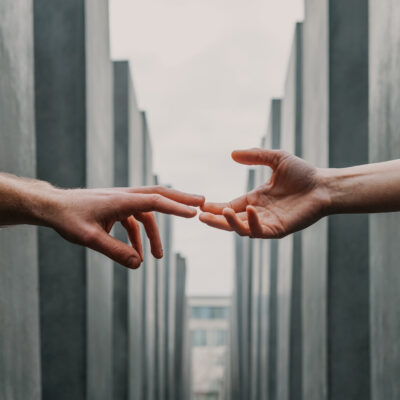Au premier plan, deux mains essaient de s'atteindre. Elles se touchent presque du bout des doigts. L'arrière-plan est fait de différents tons de gris. On peut y voir comme ligne de fuite un couloir de piliers faits de ciment, rectangulaires et imposants. Cela laisse penser à une allée du mémorial de l'holocauste à Berlin.