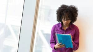 Une femme en chemise de couleur violette travaille debout, au bord d’une fenêtre, sur une tablette. Elle est face à nous, concentrée sur son travail. Illustration du live autour du livre et de l’émission : Tous hétéros au boulot ?, du podcast Méta de Choc.