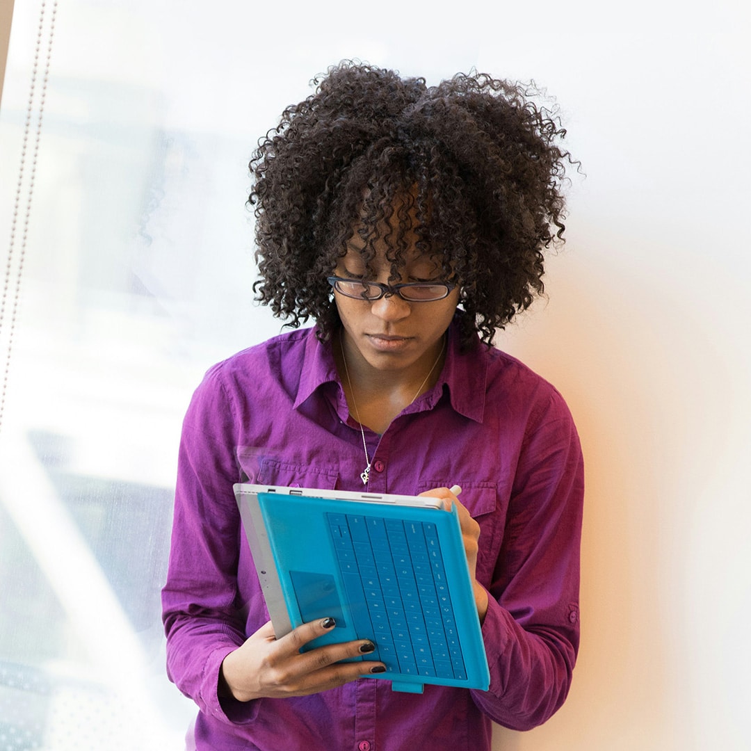 Une femme en chemise de couleur violette travaille debout, au bord d’une fenêtre, sur une tablette. Elle est face à nous, concentrée sur son travail. Illustration du live autour du livre et de l’émission : Tous hétéros au boulot ?, du podcast Méta de Choc.