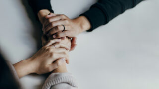 Deux paires de mains se tiennent et s'enlacent sur une table blanche. Illustration du teaser de l’émission STREAM intitulée : Santé mentale : fausses promesses et solutions, du podcast Méta de Choc.