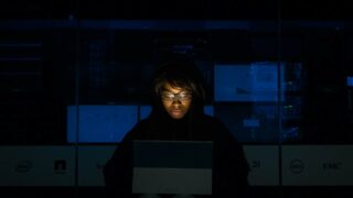 Une femme portant des lunettes regarde un ordinateur portable éclairé dans une pièce sombre. Illustration du teaser de l’émission STREAM intitulée : Formations bullshit : comment les éviter ?, du podcast Méta de Choc.